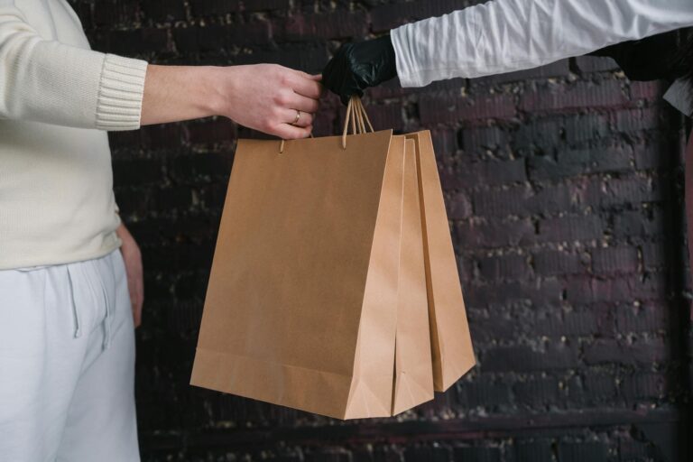 Two individuals exchange paper shopping bags against a dark brick wall background.